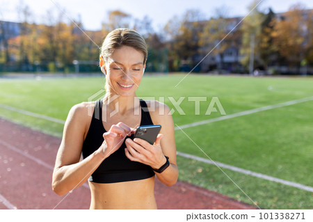 Young sporty woman athlete and runner standing smiling in stadium in sportswear and using phone. Young sporty woman athlete and runner standing smiling in stadium in sportswear and using phone. 101338271