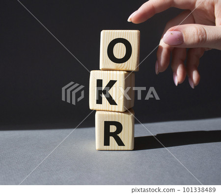 OKR objectives and key results symbol. Wooden cubes with words OKR objectives and key results. Businessman hand. Beautiful grey background. Business and OKR concept. Copy space. OKR objectives and key results symbol. Wooden cubes with words OKR objectives and key results. Businessman hand. Beautiful grey background. Business and OKR concept. Copy space. 101338489