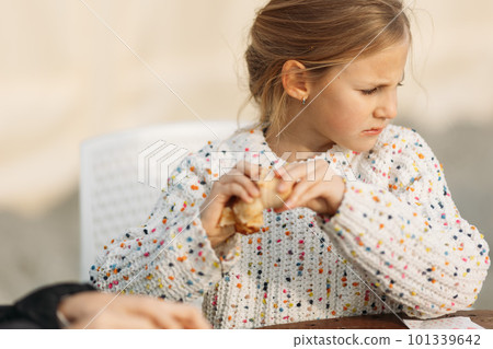 Serious little girl in white pullover sitting in cafe outdoors, eating her pancake and listening to her friend Serious little girl in white pullover sitting in cafe outdoors, eating her pancake and listening to her friend 101339642