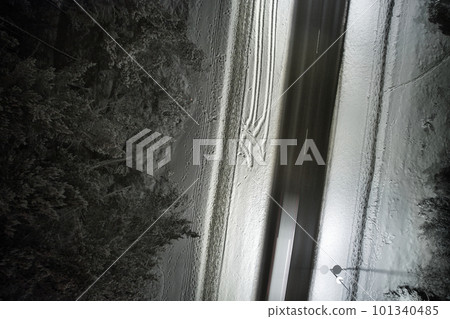 An atmospheric drone photo of a winter road winding through a snow-covered forest at night. The trees are adorned with a delicate layer of hoarfrost, creating a magical winter wonderland 101340485