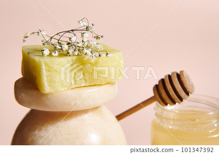 Selective focus on a bar of cold-pressed homemade natural organic soap with floral and herbal ingredients, displayed on a beige marble surface against a jar with honey, on pink pastel background 101347392