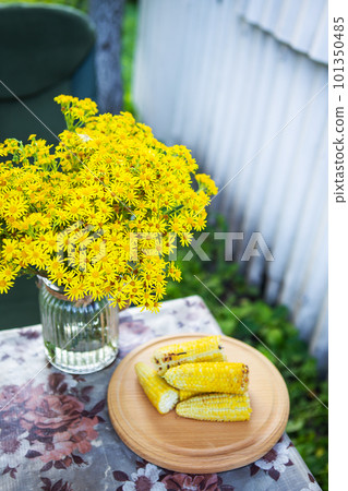 Delicious, sweet young grilled fried corn lies on a wooden board on the table along with a bouquet of flowers, outdoor recreation. Delicious, sweet young grilled fried corn lies on a wooden board on the table along with a bouquet of flowers, outdoor recreation. 101350485