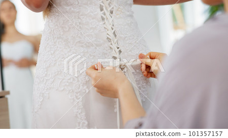 Friends helping woman with wedding dress knot in a room or boutique for marriage ready, fitting or fashion with corset lace detail and hands. Bride with mirror reflection getting ready for love event Friends helping woman with wedding dress knot in a room or boutique for marriage ready, fitting or fashion with corset lace detail and hands. Bride with mirror reflection getting ready for love event 101357157