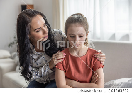 Mother comforts upset daughter that sits with arms crossed on a couch. 101363732