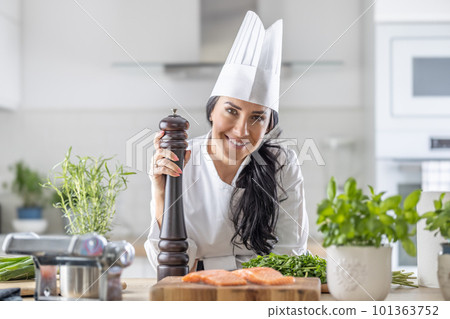 Female chef in toque blanche, white hat, and uniform holds a pepper grinder during fish and veg meal preparation Female chef in toque blanche, white hat, and uniform holds a pepper grinder during fish and veg meal preparation 101363752