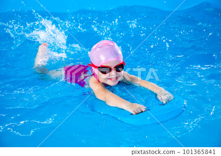 Smiling little girl learning to swim in indoor pool with flutterboard during swimming class Smiling little girl learning to swim in indoor pool with flutterboard during swimming class 101365841
