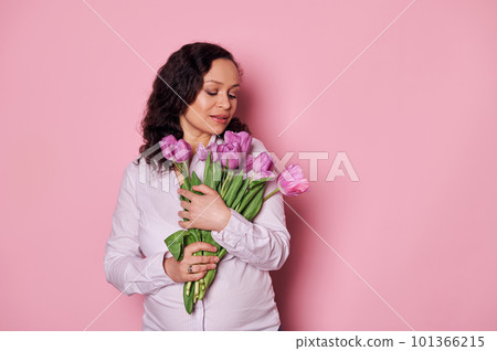 Delightful wavy-haired brunette, middle-aged pregnant woman holding bunch of purple tulips, isolated on pink background. Spring and femininity concept. Happy Mother's Day. Portrait of beautiful women Delightful wavy-haired brunette, middle-aged pregnant woman holding bunch of purple tulips, isolated on pink background. Spring and femininity concept. Happy Mother's Day. Portrait of beautiful women 101366215