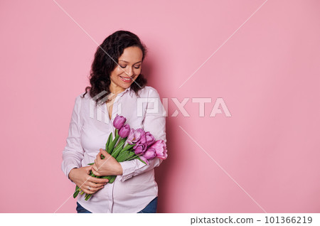 Beautiful young pregnant woman feeling happy, receiving a cute bouquet of purple tulips, smiles and expresses positive emotions, isolated over pink color background. Mother's Day and maternity concept Beautiful young pregnant woman feeling happy, receiving a cute bouquet of purple tulips, smiles and expresses positive emotions, isolated over pink color background. Mother's Day and maternity concept 101366219