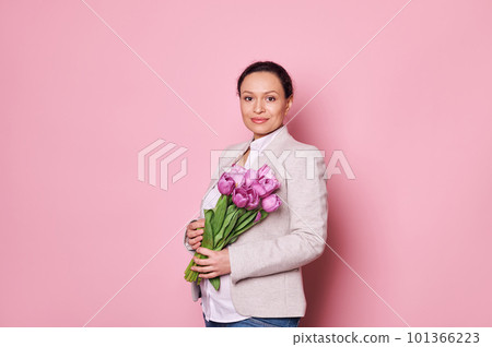 Portrait of a beautiful pregnant middle-aged woman in casual clothes, holding a bouquet of purple tulips, smiling at camera, isolated on pink background. Happy Mother's Day. International Women's Day Portrait of a beautiful pregnant middle-aged woman in casual clothes, holding a bouquet of purple tulips, smiling at camera, isolated on pink background. Happy Mother's Day. International Women's Day 101366223