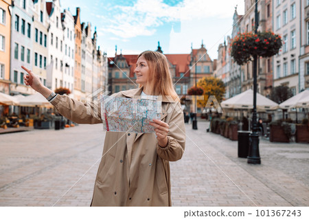 Happy woman tourist pointing finger and holding a paper map, searching direction on location map during vacation in Gdansk old town. Happy woman tourist pointing finger and holding a paper map, searching direction on location map during vacation in Gdansk old town. 101367243