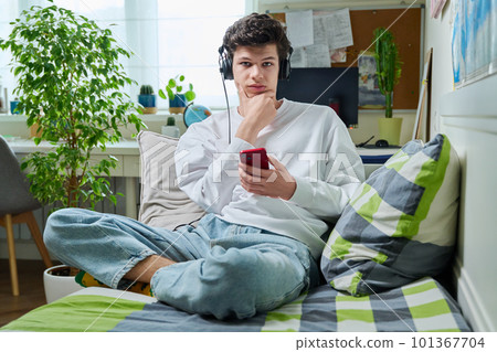 Young male in headphones with phone looking at camera sitting on couch at home Young male in headphones with phone looking at camera sitting on couch at home 101367704