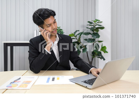 Young asian businessman in white shirt and black suit using mobile phone while look at laptop computer monitor. 101371740