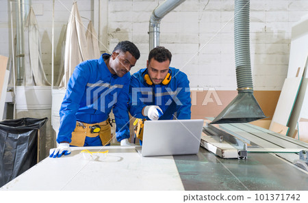 Young middle eastern labour in mechanic jumpsuit, ear muff and tool belt pointing at laptop computer monitor while working in a furniture factory. A technician stand beside him. 101371742