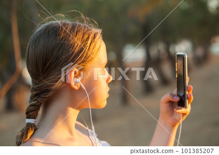 Teenage girl taking picture of sunset on mobile phone against forest background 101385207