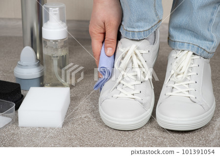 Cleaning white shoes, caring for sneakers. A woman wipes her leather shoes with a wet cloth from dust and dirt. 101391054