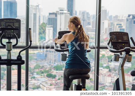 Young woman on a stationary bike in a gym on a big city background Young woman on a stationary bike in a gym on a big city background 101391305