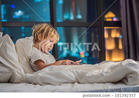 The boy uses the tablet in his bed before going to sleep on a background of a night city. Children and technology concepts 101392357
