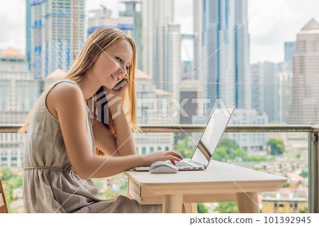 Young woman is working on a laptop on her balcony overlooking the skyscrapers. Freelancer, remote work, work from home Young woman is working on a laptop on her balcony overlooking the skyscrapers. Freelancer, remote work, work from home 101392945