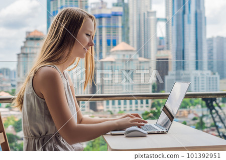 Young woman is working on a laptop on her balcony overlooking the skyscrapers. Freelancer, remote work, work from home Young woman is working on a laptop on her balcony overlooking the skyscrapers. Freelancer, remote work, work from home 101392951
