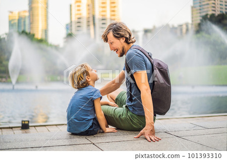 Dad and son tourists on background of fountain on the lake at night, near by Twin Towers with city on background. Kuala Lumpur, Malaysia Dad and son tourists on background of fountain on the lake at night, near by Twin Towers with city on background. Kuala Lumpur, Malaysia 101393310