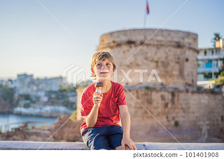 Boy tourist eating turkish ice cream on background of Hidirlik Tower in Antalya against the backdrop of the Mediterranean bay of the ancient Kaleici district, Turkey. Turkiye 101401908