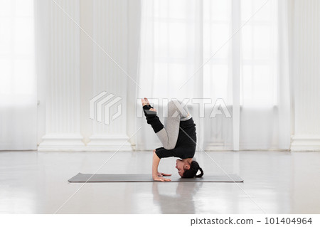 Side view of an athletic woman doing headstand training during yoga meditation in a bright gym. Concept of normalization of all processes in the body, strength, energy and concentration 101404964