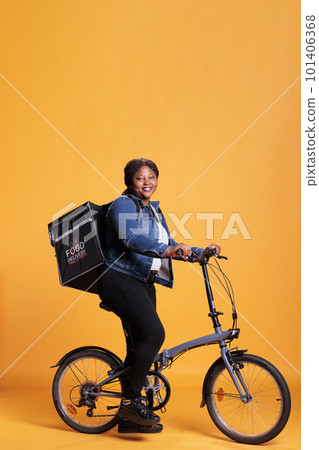 Pizzeria worker carrying food delivery thermal backpack standing riding bike in studio with yellow background. Restaurant courier delivering pizza for lunch. Food transportation service 101406368