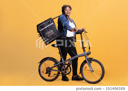 Portrait of restaurant worker standing beside bike holding takeaway delivery thermal backpack, delivering fast food order to customer. Pizzeria courier standing in studio over yellow background Portrait of restaurant worker standing beside bike holding takeaway delivery thermal backpack, delivering fast food order to customer. Pizzeria courier standing in studio over yellow background 101406389