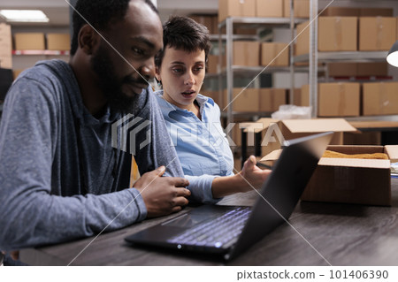 Diverse employees sitting at table in warehouse checking transportation logistics on laptop computer before delivery trendy blouse. Fashion retail workers putting clothes in cardboard box Diverse employees sitting at table in warehouse checking transportation logistics on laptop computer before delivery trendy blouse. Fashion retail workers putting clothes in cardboard box 101406390
