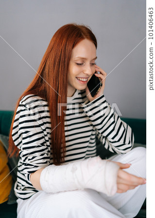 Vertical medium shot of happy young woman injured hand wrapped in gypsum bandage talking on smartphone with friends during rehabilitation. Smiling female with with broken arm from accident at home. 101406463