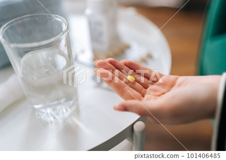 Close-up cropped shot of unrecognizable young woman holding pill taking medicine for flu, influenza, painkiller, vitamin complex, oral contraceptives on palm on blurred background of table with drugs. Close-up cropped shot of unrecognizable young woman holding pill taking medicine for flu, influenza, painkiller, vitamin complex, oral contraceptives on palm on blurred background of table with drugs. 101406485