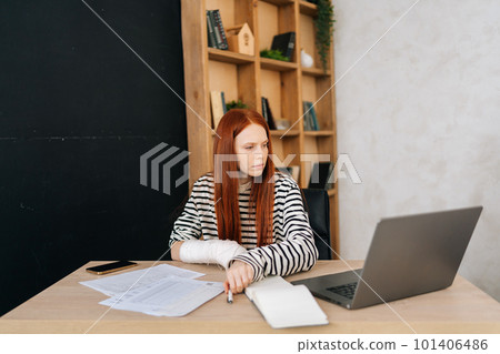 Serious female freelancer with broken right hand wrapped in white gypsum bandage working on laptop computer sitting at table. Portrait of injured businesswoman working remotely from home office. Serious female freelancer with broken right hand wrapped in white gypsum bandage working on laptop computer sitting at table. Portrait of injured businesswoman working remotely from home office. 101406486