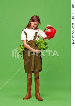 Shot of young pretty smiling girl, gardener watering plants from a watering can over green background. Takes care of nature 101407304