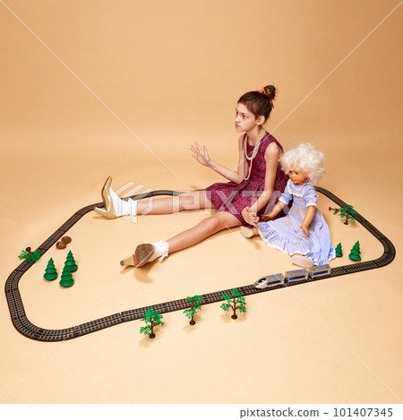 Portrait of emotionless little girl wearing dress and heels sitting with doll over beige background. Neutral emotions Portrait of emotionless little girl wearing dress and heels sitting with doll over beige background. Neutral emotions 101407345
