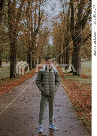 a guy in a jacket stands on an alley in the park during the fall season. Portrait oh handsome teen guy, young man in hoodie, down jacket standing, walking in beautiful golden autumn park, looking at 101409174