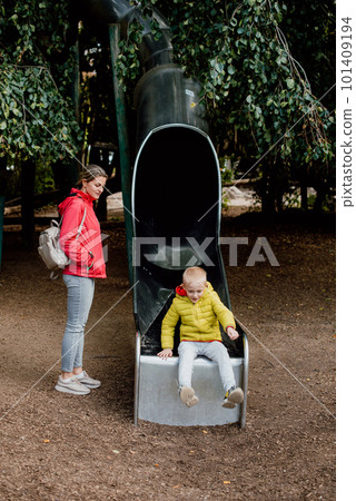 Curious toddler boy in warm clothes playing on the playground slide with his mommy. Caucasian mother stand and take care little boy play slide in fun park of garden and they look happy to have enjoy Curious toddler boy in warm clothes playing on the playground slide with his mommy. Caucasian mother stand and take care little boy play slide in fun park of garden and they look happy to have enjoy 101409194