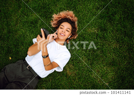 Young woman sitting on the grass in a city park and using a smartphone. Technology and modern lifestyle concept. 101411141