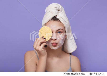 Horizontal shot of young attractive girl posing with white towel on head, holding sponge in hand and covering her eye with it, keeping lips rounded, making kiss gesture, morning cosmetic procedures. 101411233