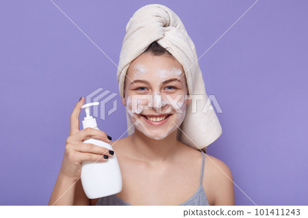 Horizontal indoor picture of adorable sincere sweet female looking directly at camera, holding bottle with cleansing agent, applying facial mask, having peaceful facial expression. Skin care concept. 101411243