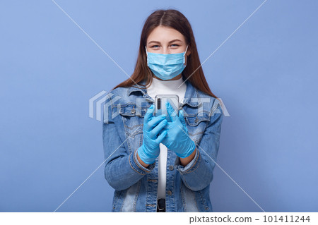 Portrait of happy magnetic attractive young female in antibacterial mask and gloves, holding smartphone in hands, being isolated, using device, being in quarantine. Coronavirus outbreak concept. 101411244