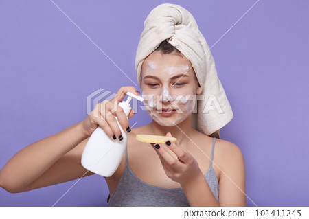 Indoor shot of young woman applying cream on sponge while posing isolated over lilac background, female looking concentrated on sponge with gel or lotion, doing morning cosmetic procedures at home. 101411245