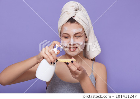 Indoor shot of young charming woman wrapped white towel using cleansing foam from dispenser, applies agent on cosmetic sponge, doing morning procedures, posing isolated over lilac background. Indoor shot of young charming woman wrapped white towel using cleansing foam from dispenser, applies agent on cosmetic sponge, doing morning procedures, posing isolated over lilac background. 101411247