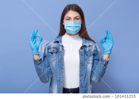 Picture of young woman showing sign ok, invoking to not panic, lady wearing protective face mask and disposable medical gloves, preventing virus infection, isolated over blue background. Coronavirus Picture of young woman showing sign ok, invoking to not panic, lady wearing protective face mask and disposable medical gloves, preventing virus infection, isolated over blue background. Coronavirus 101411255