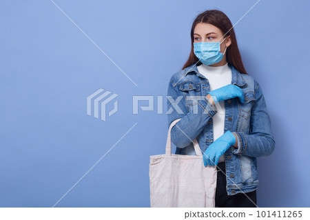 Picture of attentive sweet model with long hair looking aside, being in mask and gloves for safety, coronavirus outbreak, keeping distance in quarantine while shopping. Copyspace for advertisement. 101411265