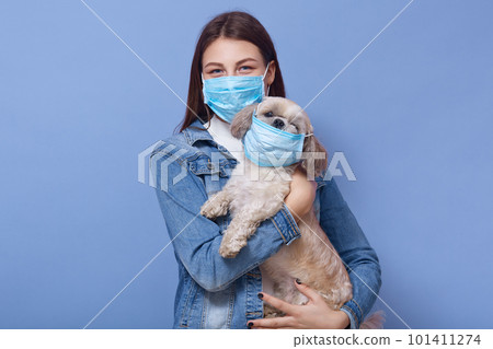 Studio shot of young dark haired woman wearing medical face mask standing together with her dog dresses protective mask too, isolated over blue background. Coronavirus, covid 19, health care concept. 101411274