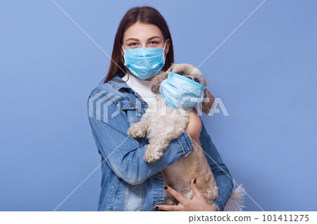 Woman wearing denim jacket and protective medical mask holding dog pet in face flumask, posing isolated over blue studio background. Coronavirus disease COVID-19, dangerous for people and pets. 101411275