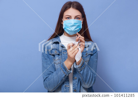 Indoor shot of careful young female standing isolated over blue background, wiping her hands with antibacterial tissue, killing germs, sticking to safety measures, being in mask. Coronavirus concept. Indoor shot of careful young female standing isolated over blue background, wiping her hands with antibacterial tissue, killing germs, sticking to safety measures, being in mask. Coronavirus concept. 101411285
