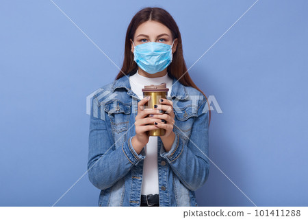 Pictire of young Caucasian woman waring denim jacket and disposable medical mask, holding coffee or tea in thermo mug, posing isolated over blue studio background. Coronavirus, covid 19 concept. Pictire of young Caucasian woman waring denim jacket and disposable medical mask, holding coffee or tea in thermo mug, posing isolated over blue studio background. Coronavirus, covid 19 concept. 101411288