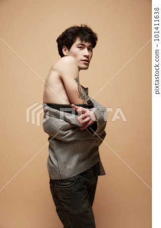 Portrait of young asian man with tattoo on hand posing shirtless in stylish grey jacket against light brown studio background Portrait of young asian man with tattoo on hand posing shirtless in stylish grey jacket against light brown studio background 101411638