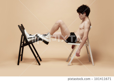 Young asian guy in underwear and stylish boots, posing shirtless on chair against light brown studio background 101411671